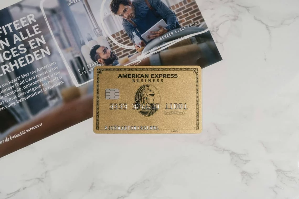 OptBlue merchant fees — AmEx Business Gold card with company brochure on marble surface, the visual register of how AmEx pitches itself to small businesses before the OptBlue fee math arrives