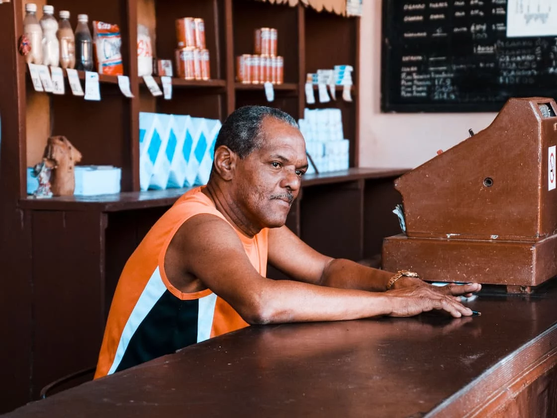 Corner-store owner at his counter beside an old register, holding the inherited merchant contract left behind by the previous owner